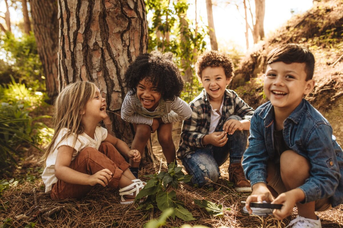 Four young children laughing and playing together in a wooded outdoor area.