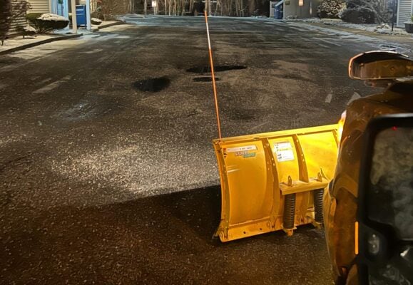 Snow plow attachment on vehicle clearing residential driveway at night in Massachusetts