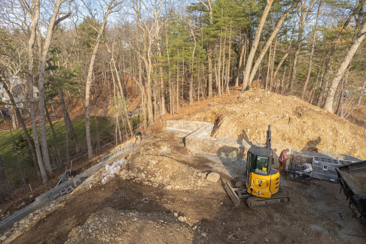 Sloped land grading project with retaining wall construction and excavator in wooded Massachusetts lot