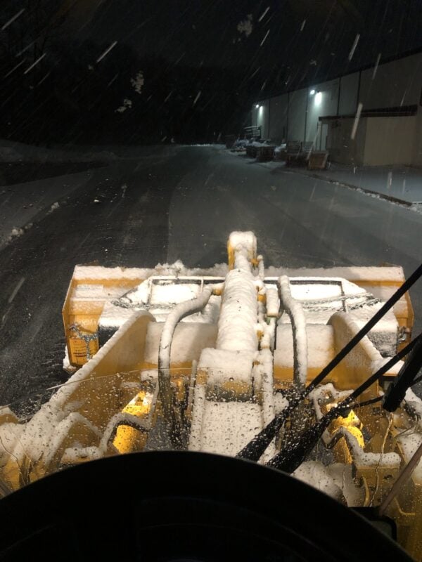 View from inside a loader cab at night, looking over the yellow hood and lift arms toward a snow-covered parking lot lit by overhead lights.