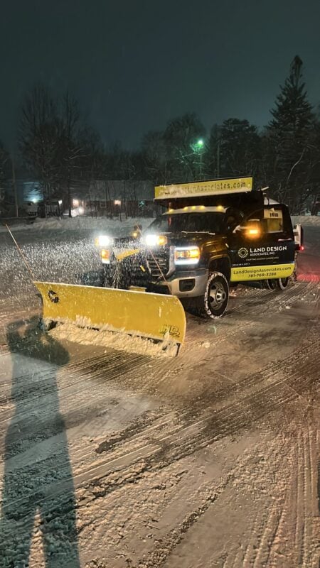 Black pickup truck with a yellow plow blade and roof light bar clearing a snow-covered parking lot at night during a snowfall