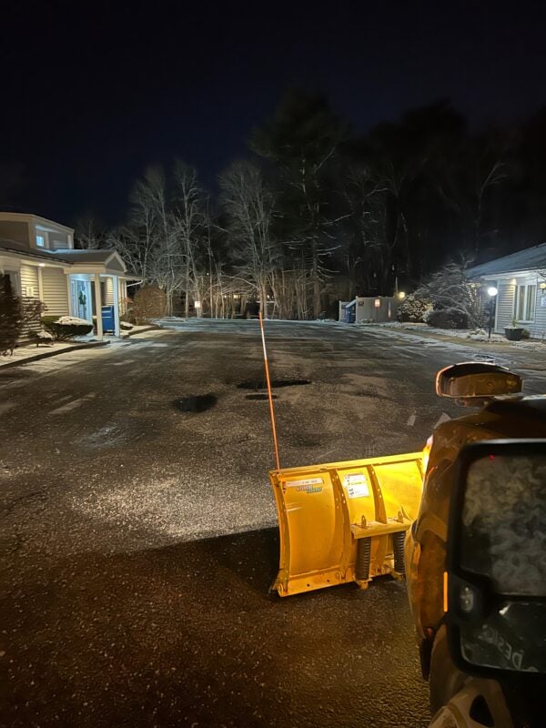 Yellow plow blade in the foreground of a freshly plowed parking lot at night, with snowbanks along the edges and lit buildings behind trees.
