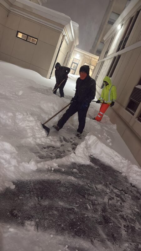Three crew members, one in a high-visibility jacket, shoveling deep snow from a walkway between buildings at night