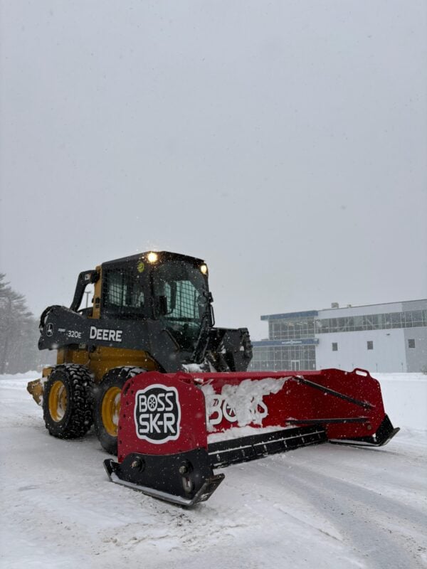 Yellow and black skid steer loader with a red snow pusher attachment clearing snow from a parking lot on an overcast day