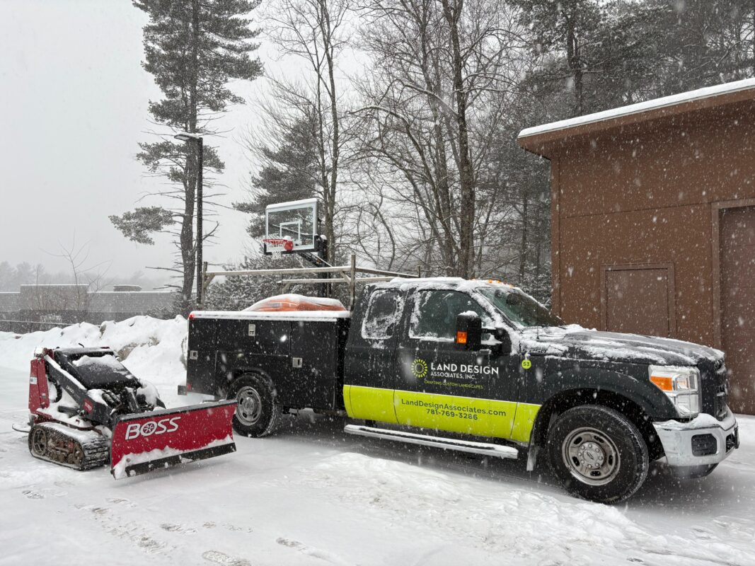 Black company pickup truck with a lime-green stripe and utility body, towing a trailer with a red compact track loader, parked in the snow beside a brick building