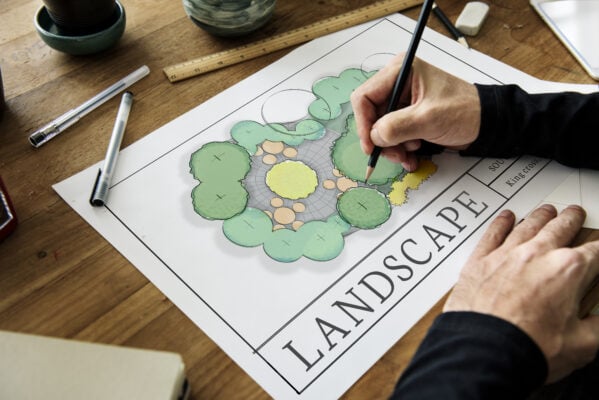 Hand drawing a colored landscape plan of a circular stone patio surrounded by trees and shrubs, on a wooden desk with a ruler and pens